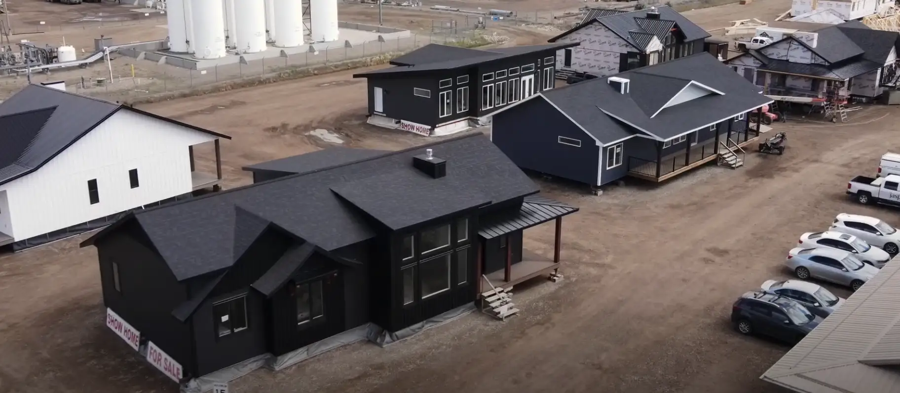 cluster of modern rtm homes in a construction yard. The homes are built in a variety of sizes and styles, featuring dark-colored exteriors, large windows, and sloping roofs. A white modular home stands out among the predominantly black structures. In the background, there are industrial storage tanks and other construction materials, indicating that this is a manufacturing or assembly site for prefabricated homes. Several cars and trucks are parked along a dirt pathway in the foreground.